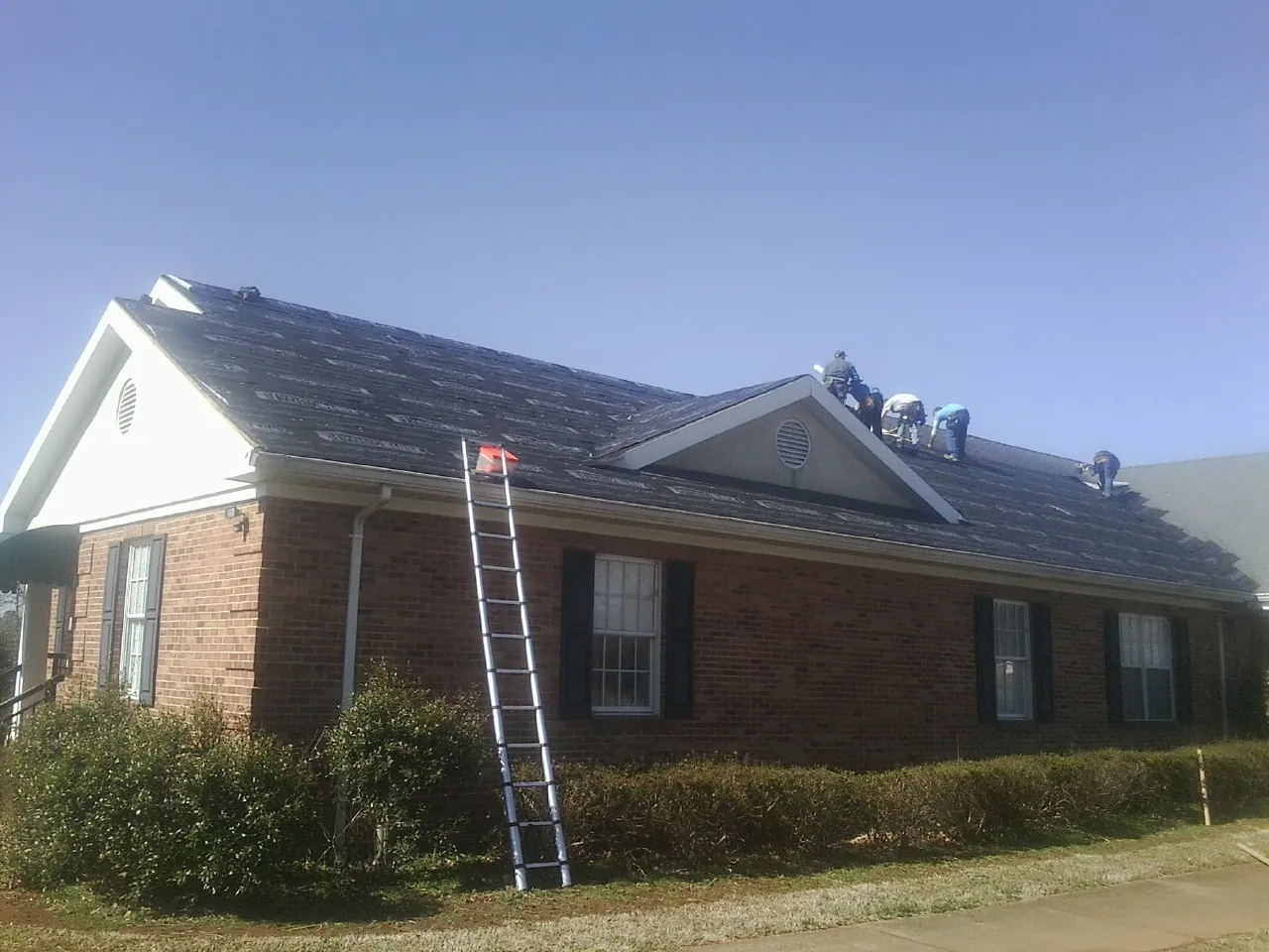 Crew tearing shingles off a brick ranch roof with a ladder set — Emergency Roof Repair work by our crew in Mc Donald